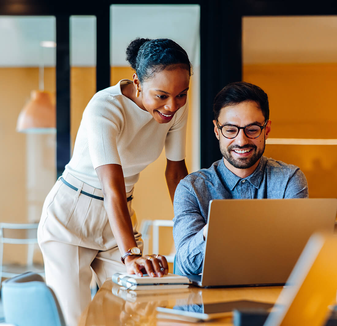 Smiling colleagues discussing project ideas while working on laptop at the office.