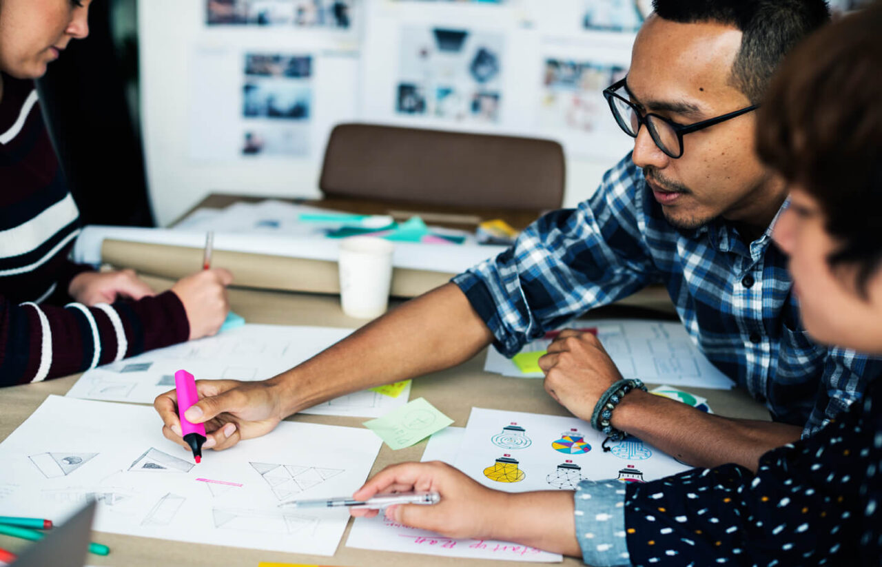Design team collaborating at a table, reviewing sketches and visual concepts with pens, markers, and notes during a creative planning session.