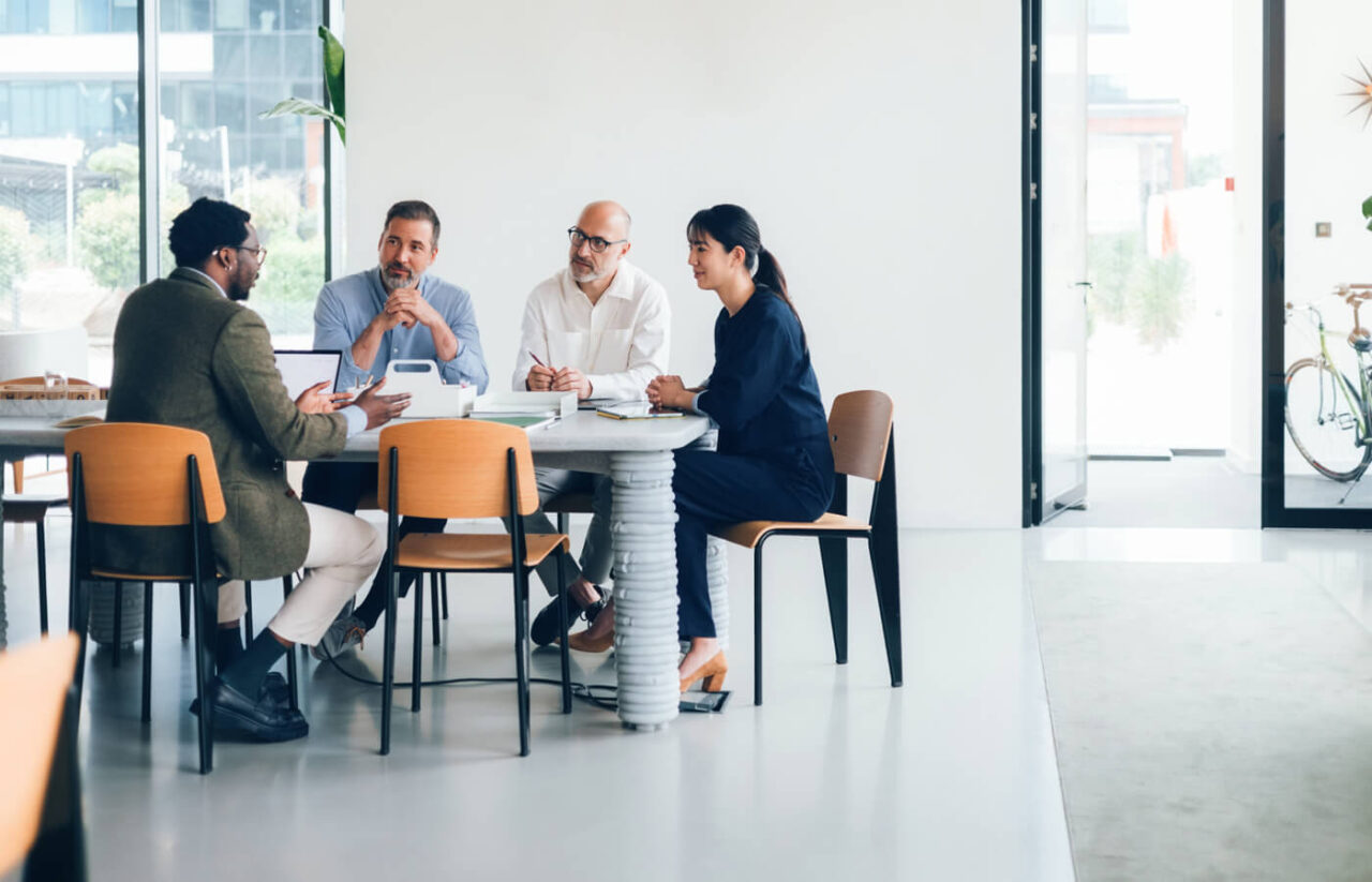 Four professionals seated around a table in a bright office, engaged in a collaborative discussion.