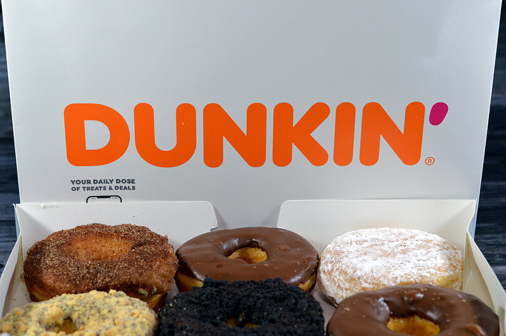 Open Dunkin' donut box displaying the modern Dunkin' wordmark above an assortment of donuts, illustrating the company's brand evolution after dropping "Donuts" from its name to reflect a broader coffee and beverage strategy.